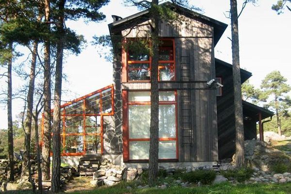 Puutalo, jossa on punakehyksiset ikkunat ja lasinen aurinkohuone metsäisessä ympäristössä.
