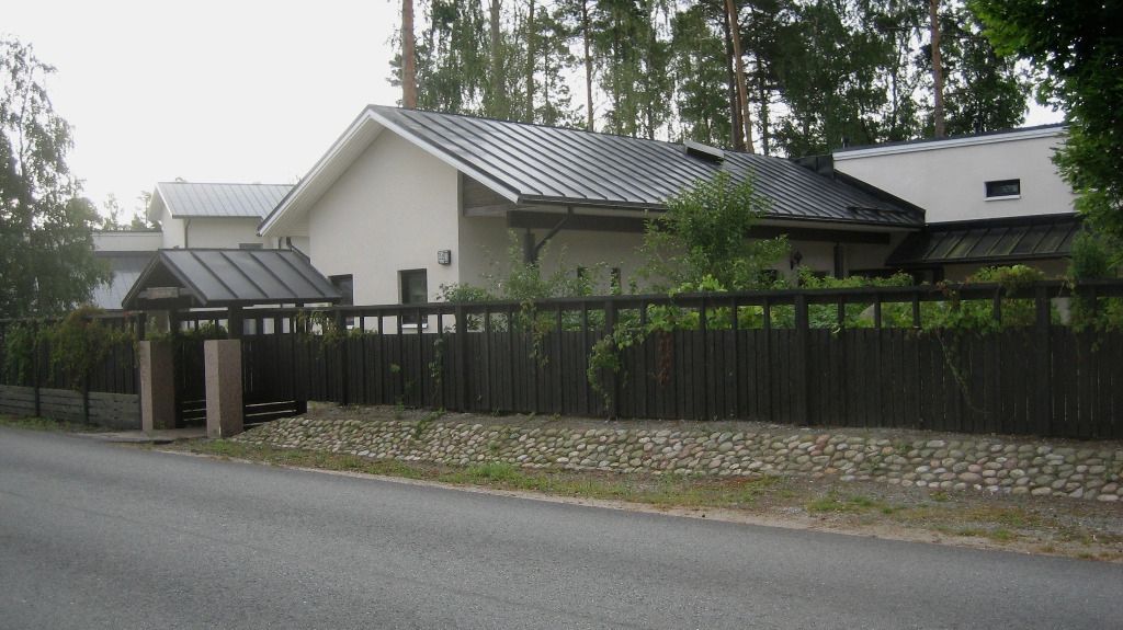Talo, jossa on tumma katto, puinen aita ja pieni portti tien varrella.