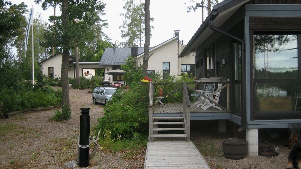 Taloja veden äärellä, puinen kävelytie ja portaat, auto ajotiellä, puita, pilvinen taivas.