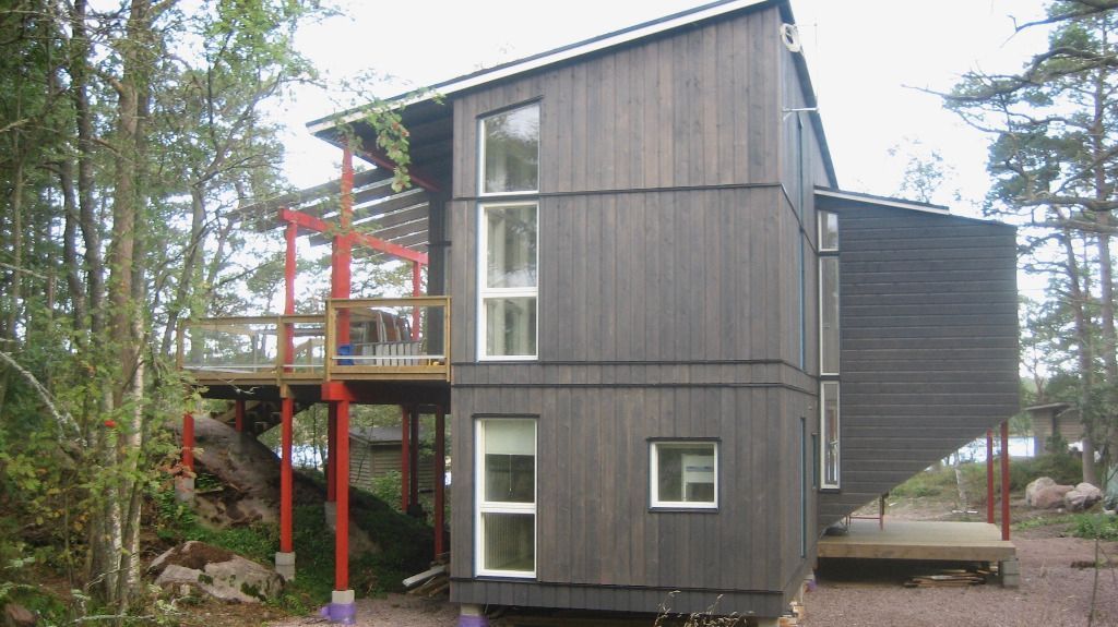 Moderni musta puutalo, jossa on punaiset tuet, terassi ja valkokehyksiset ikkunat metsäisellä alueella.