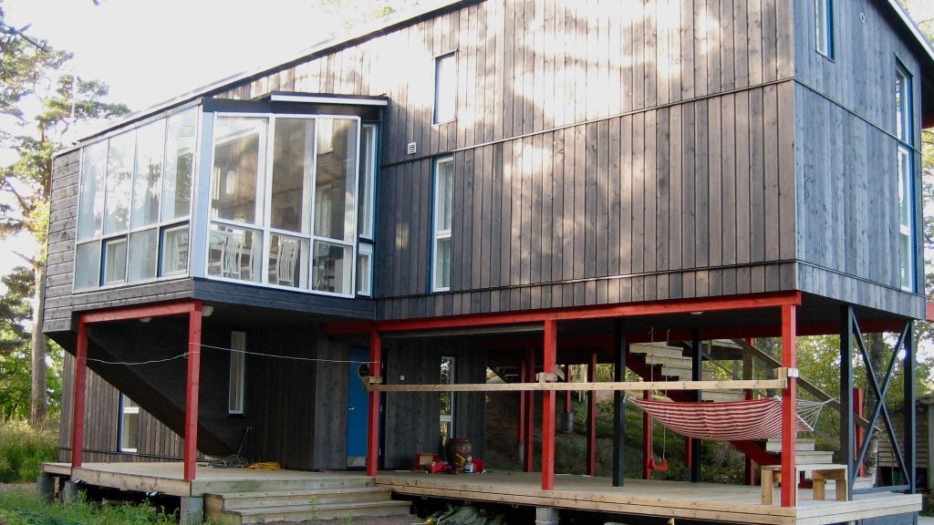 Tumma puutalo paalujen päällä, punaisilla tuilla ja lasiseinäisellä osiolla metsäisellä alueella.