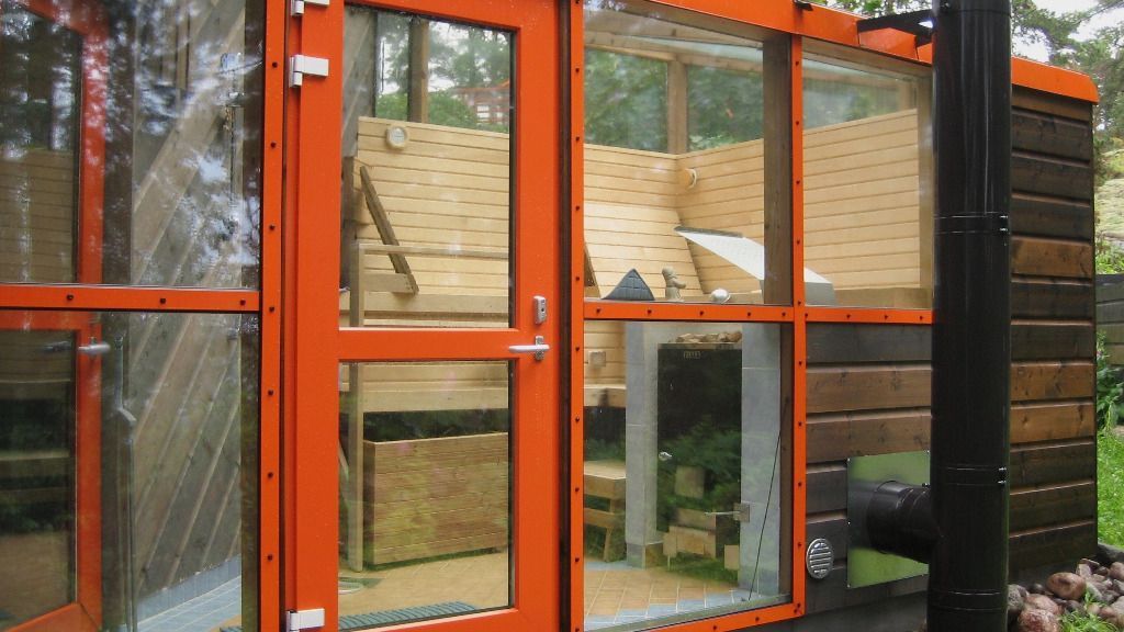 Oranssikehyksinen lasiovi ja pienen puurakenteen ikkunat, joiden sivulla on musta putki, metsäisellä alueella.