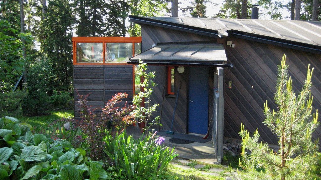 Puinen mökki, jossa on sininen ovi, oranssikehyksinen ikkuna ja jota ympäröi vihreä kasvillisuus.