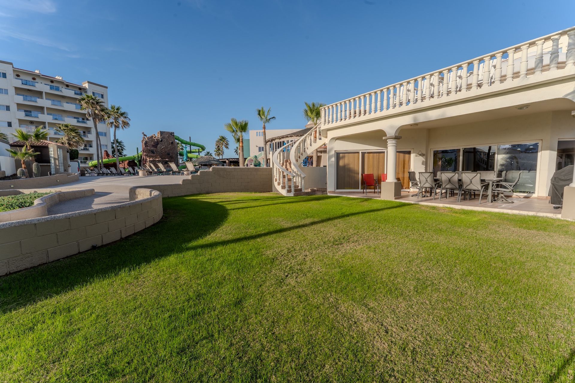 Sunny resort courtyard with green lawn, white terrace, and apartment buildings in the background