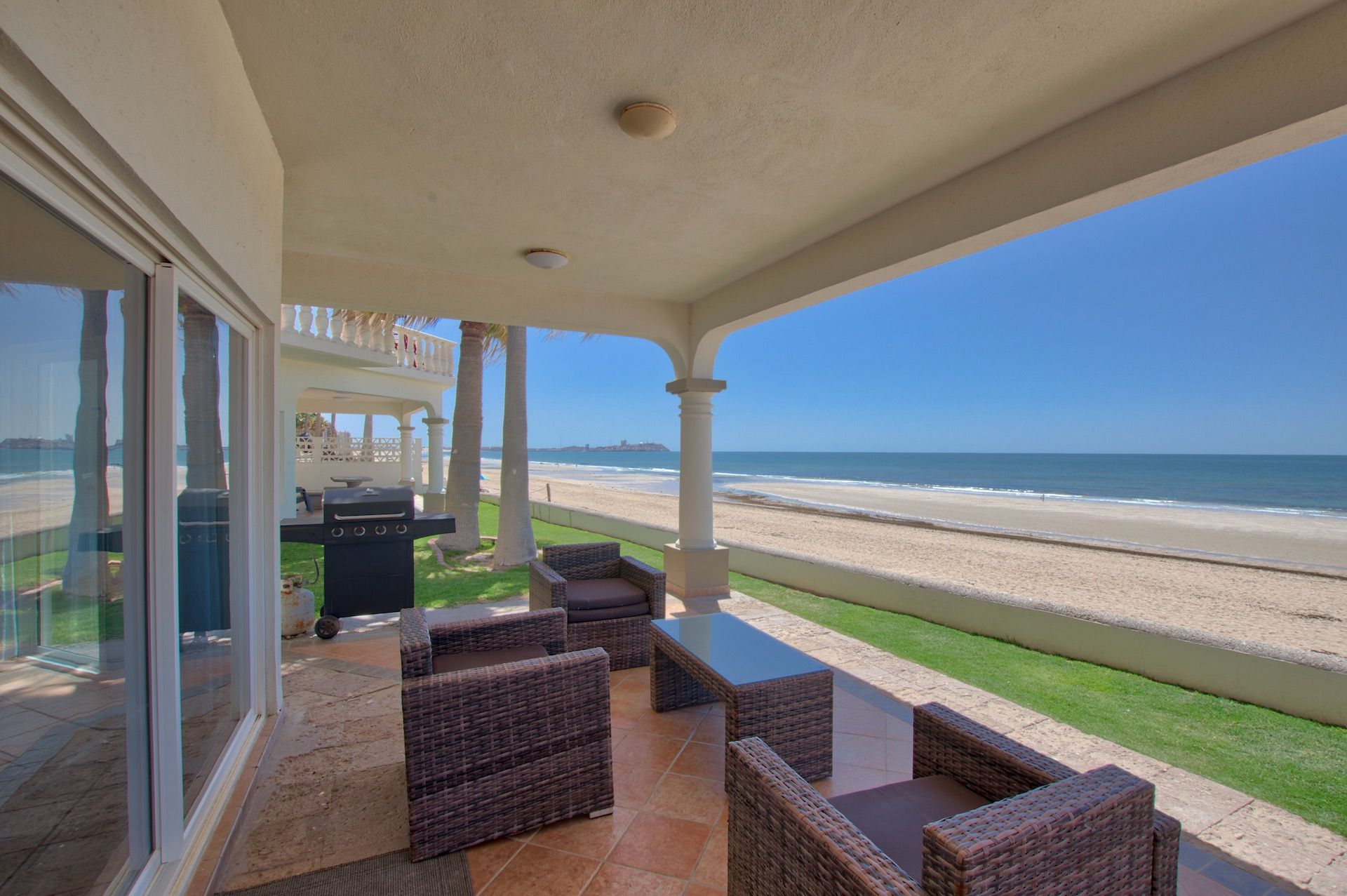 Patio overlooking a beach with seating, grill, and ocean view on a sunny day.