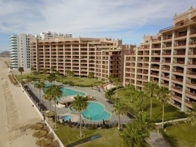 An aerial view of a tan beachside resort complex featuring multiple swimming pools, palm trees, and manicured green lawns.