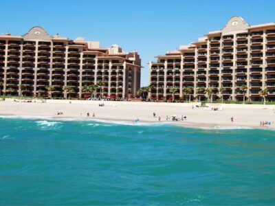 Aerial view of a curved, high-rise beach resort with an expansive pool complex and rows of umbrellas on the sand.