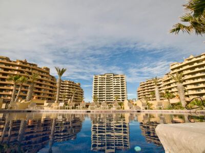 A swimming pool reflects three large, multi-story resort buildings and palm trees under a bright blue, cloudy sky.