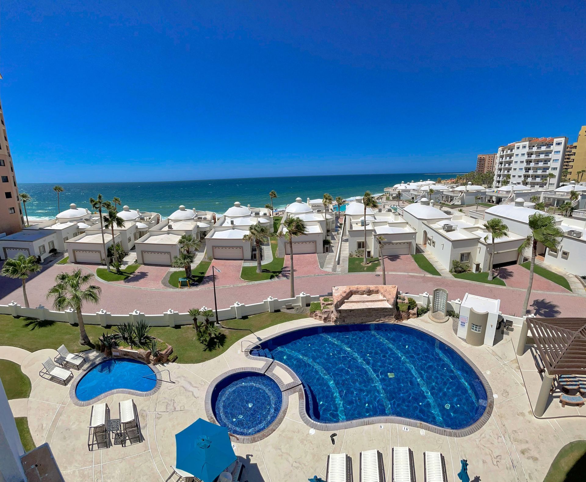 Oceanfront resort view with pool, cabanas, white buildings, and blue sky.