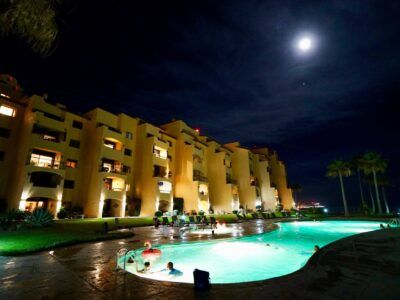 A nighttime view of a multi-story yellow resort building overlooking an illuminated swimming pool with palm trees.