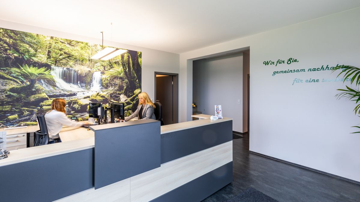 Eine Frau sitzt an einem Schreibtisch in einem Empfangsbereich. An der Wand hängt ein Bild eines Wasserfalls.