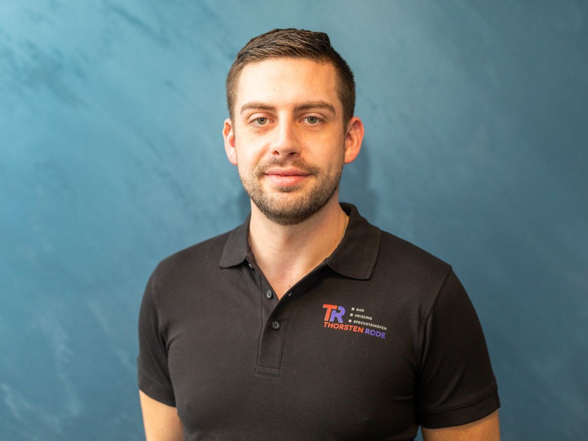 Ein Mann mit Bart und einem schwarzen Poloshirt steht vor einer blauen Wand.