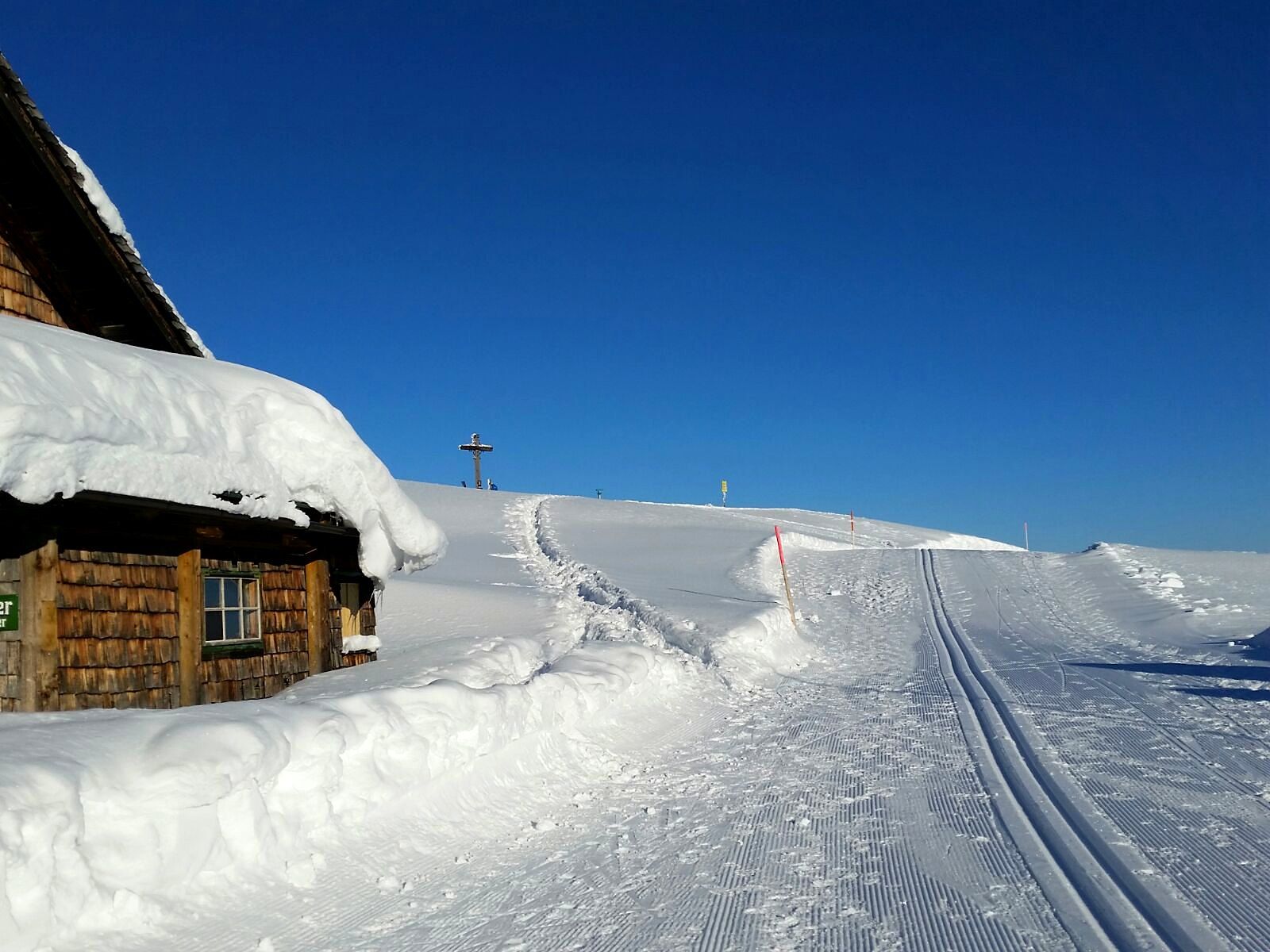 Verschneite Almhütte am Roßbrand mit Gipfelkreuz und Langlaufloipe