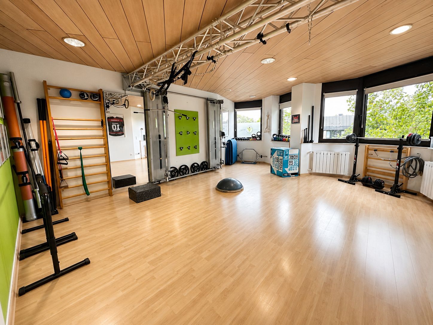 Helles Fitnessstudio mit Holzboden, Sprossenwänden, Matten und großen Fenstern.