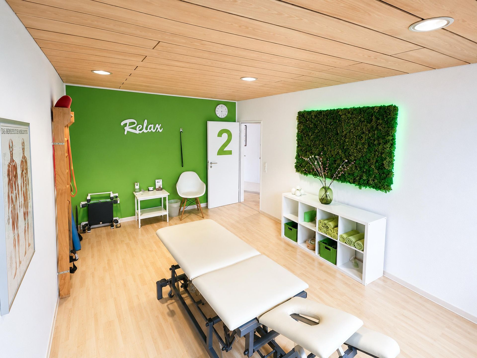 Heller Therapieraum mit grüner Akzentwand, weißem Regal, Stühlen und Pflanzen auf hellem Holzboden.