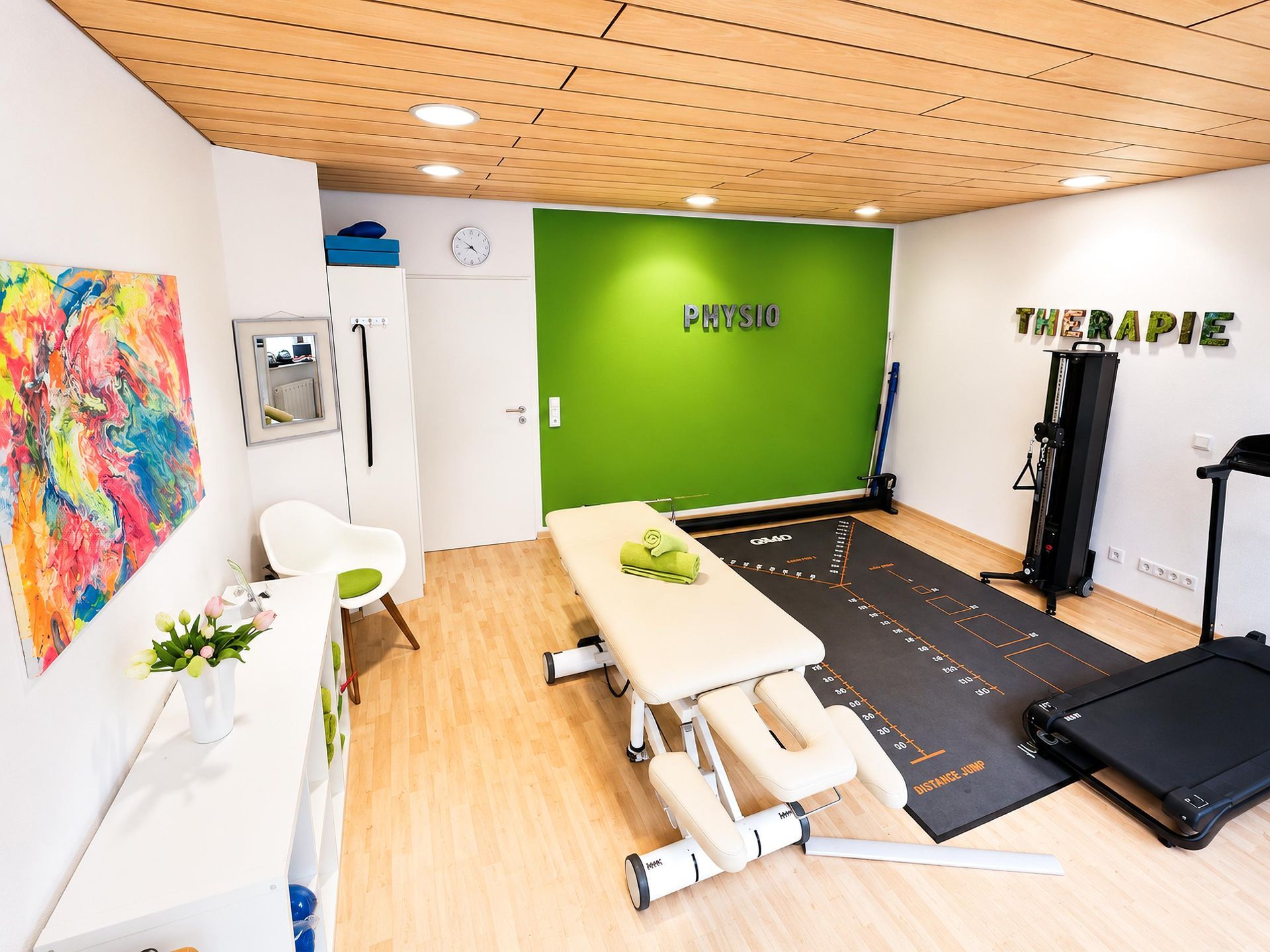 Heller Behandlungsraum mit Massageliege, grüner Akzentwand, Fitnessgeräten und farbenfroher Wandkunst.