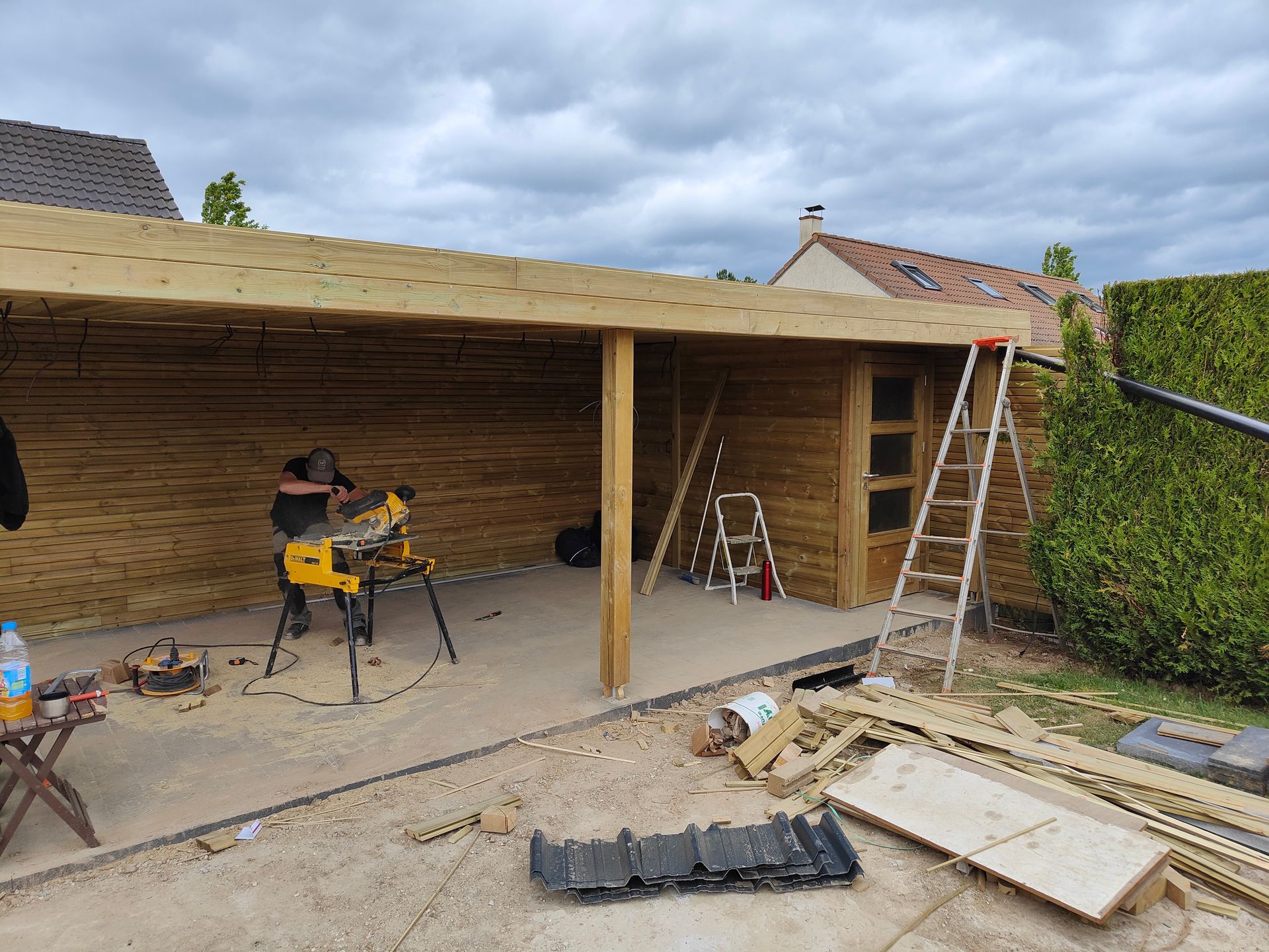 Abri de voiture en bois partiellement construit, avec des outils, des échelles et des matériaux de construction éparpillés au sol.