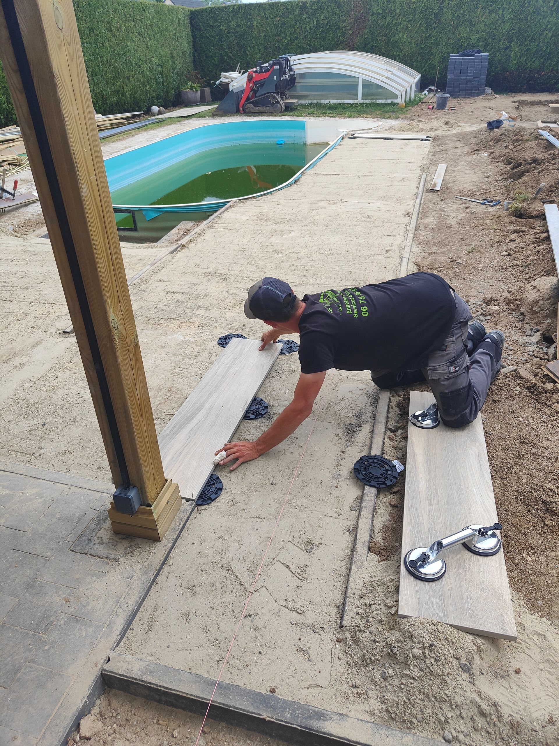 Une personne mesure un terrain sablonneux près d'une piscine, avec des planches, des outils et une clôture en arrière-plan.
