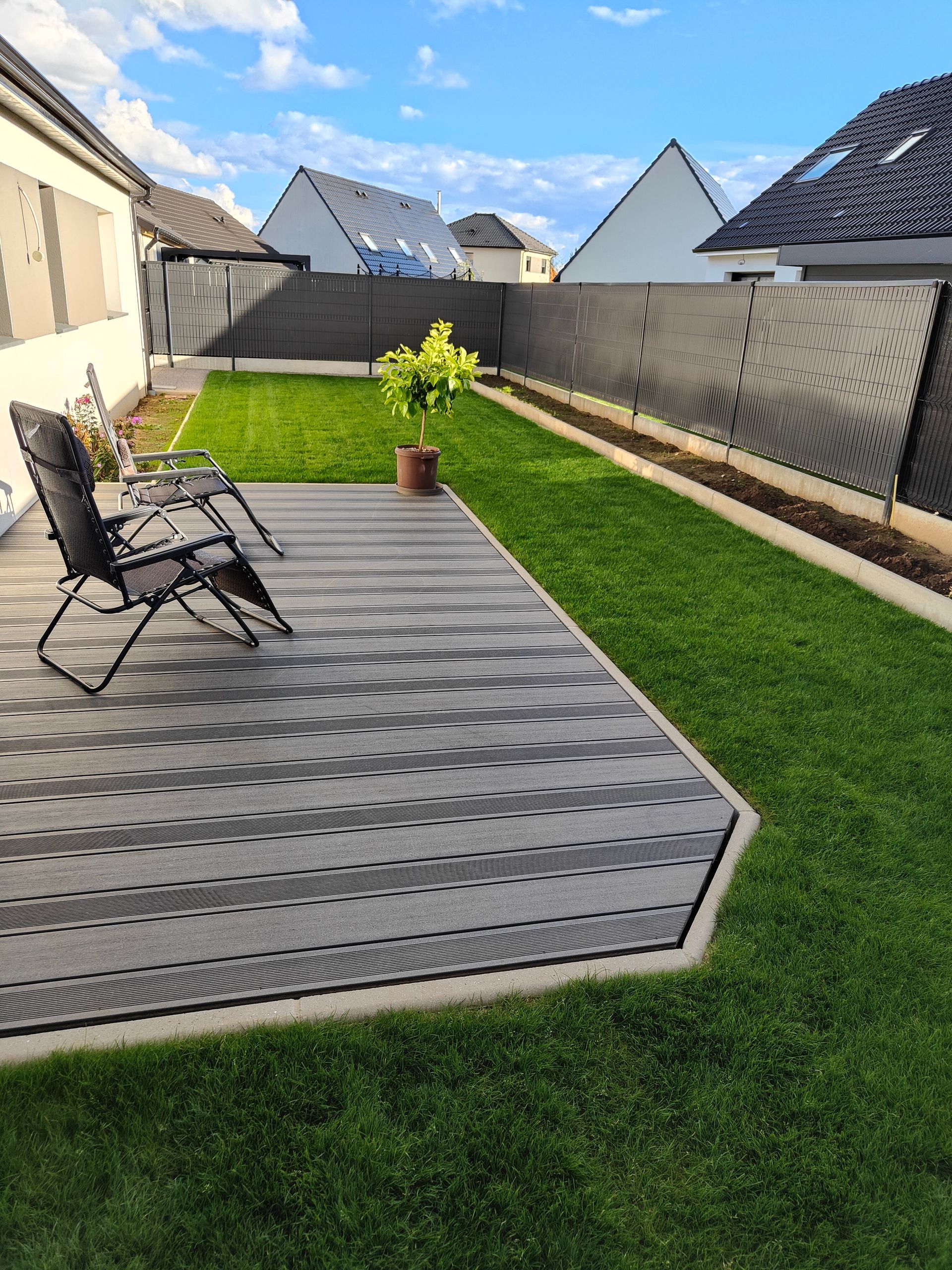 Terrasse arrière avec terrasse grise, deux chaises, plante en pot et pelouse verte sous un ciel ensoleillé