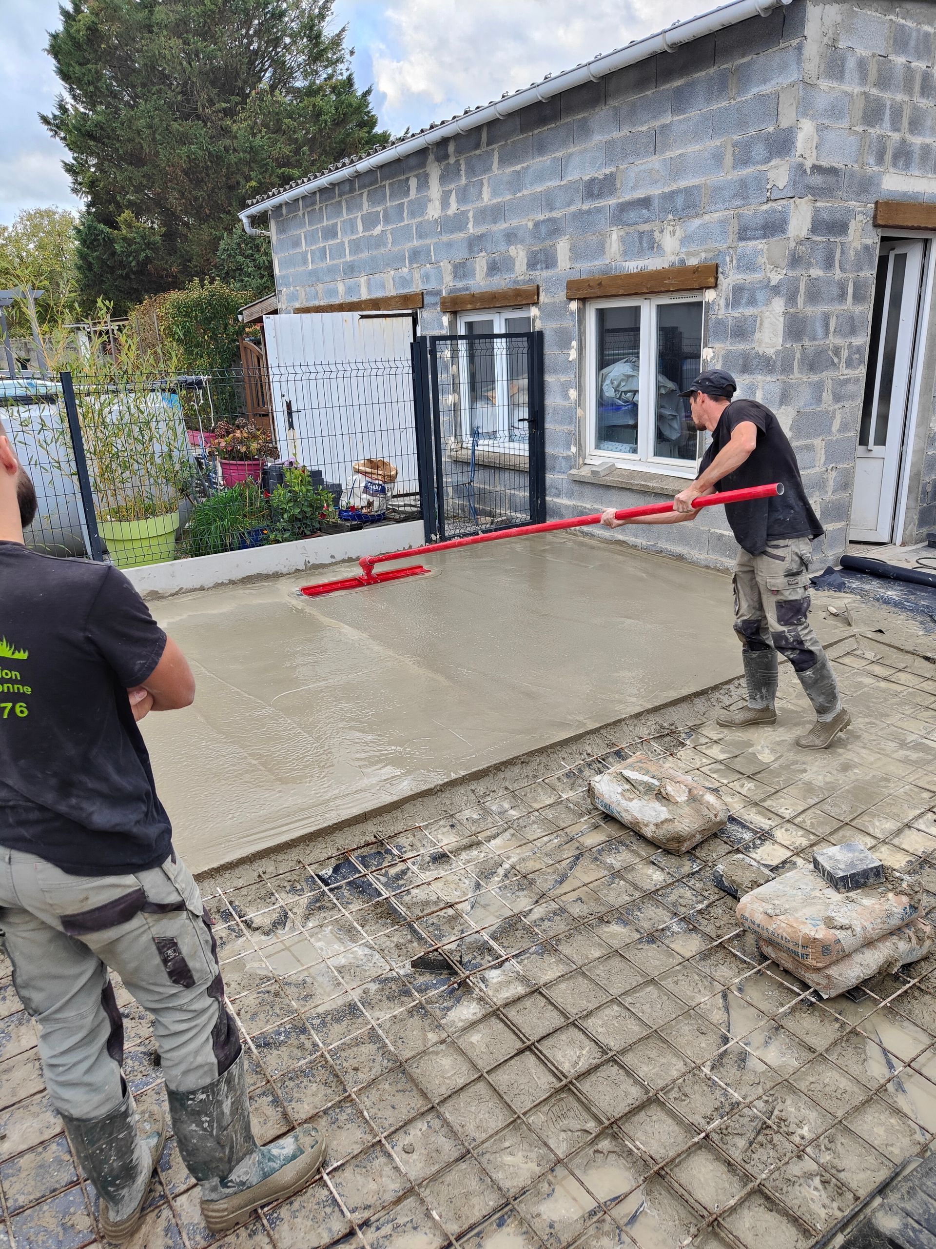 Deux ouvriers coulent du béton à côté d'un bâtiment en parpaings gris, l'un d'eux le lissant avec un outil rouge.