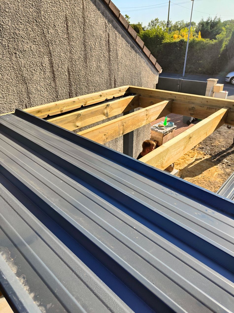 Au premier plan, des panneaux de toiture bleus et une charpente en bois apparente sur un toit en construction.