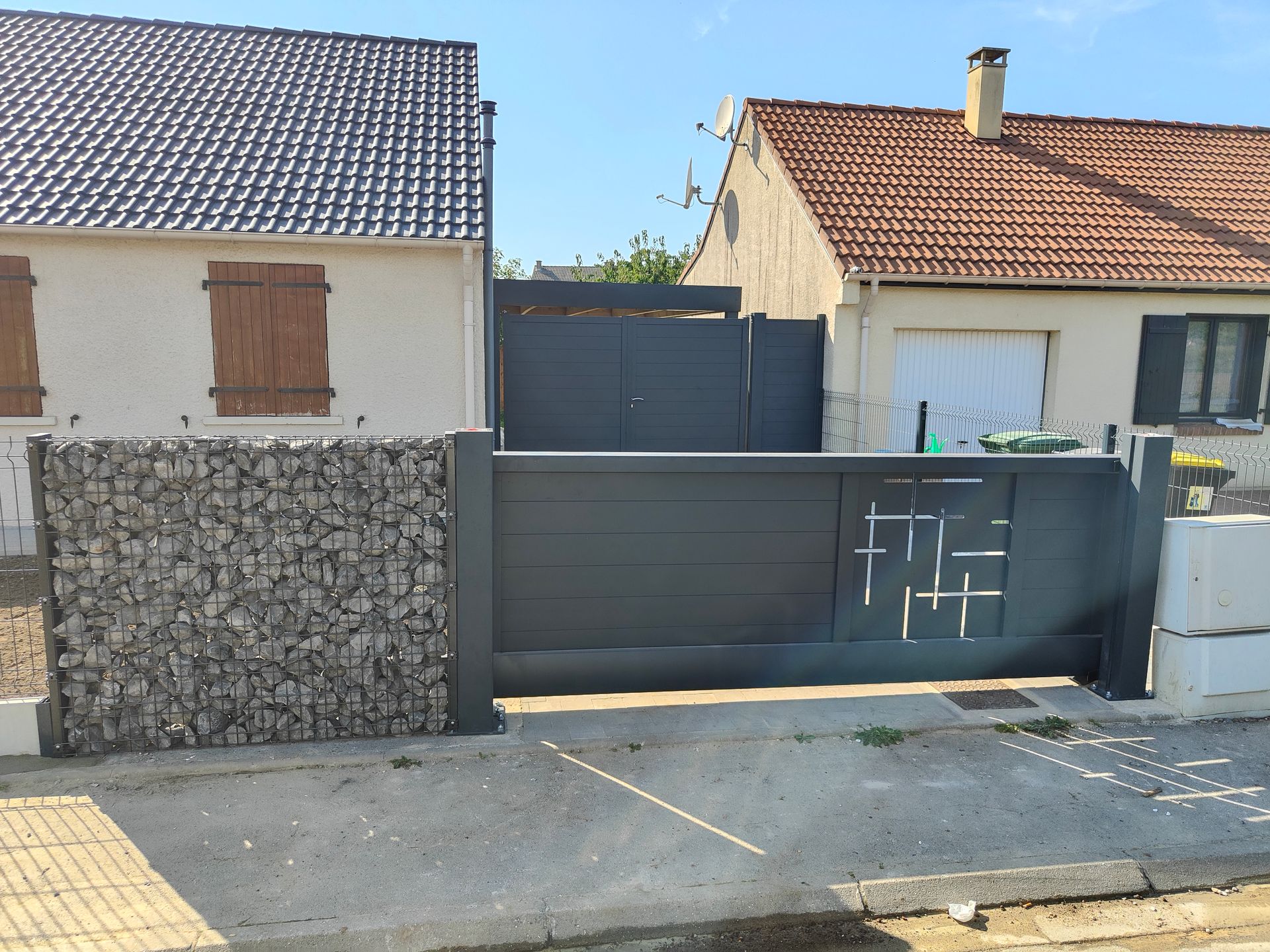 Rue résidentielle avec portail gris, mur de gabions et deux maisons aux toits de tuiles.