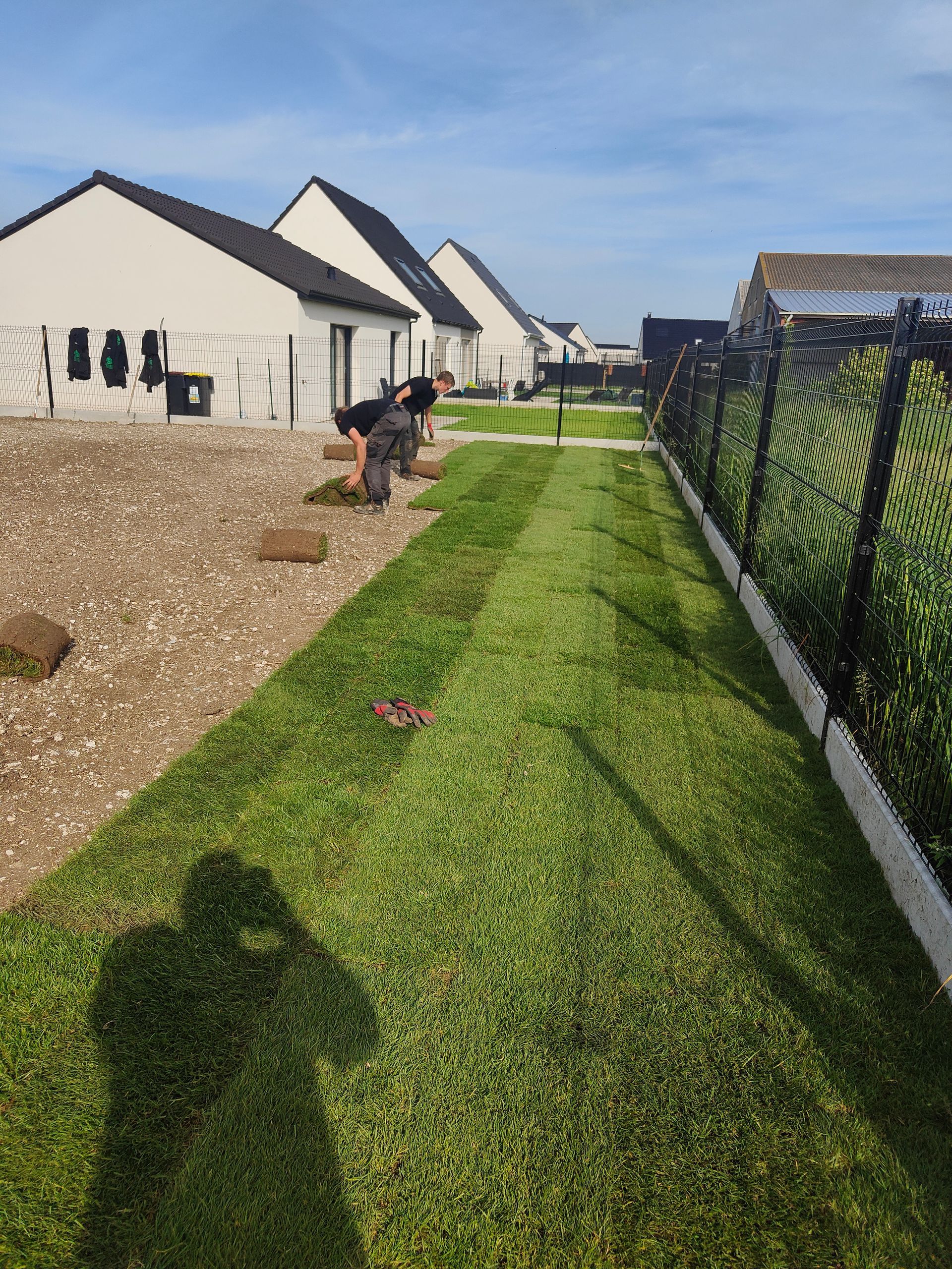 Jardin paysager avec pelouse verte, allée de gravier et maisons blanches sous un ciel bleu ; un ouvrier s'occupe du jardin.