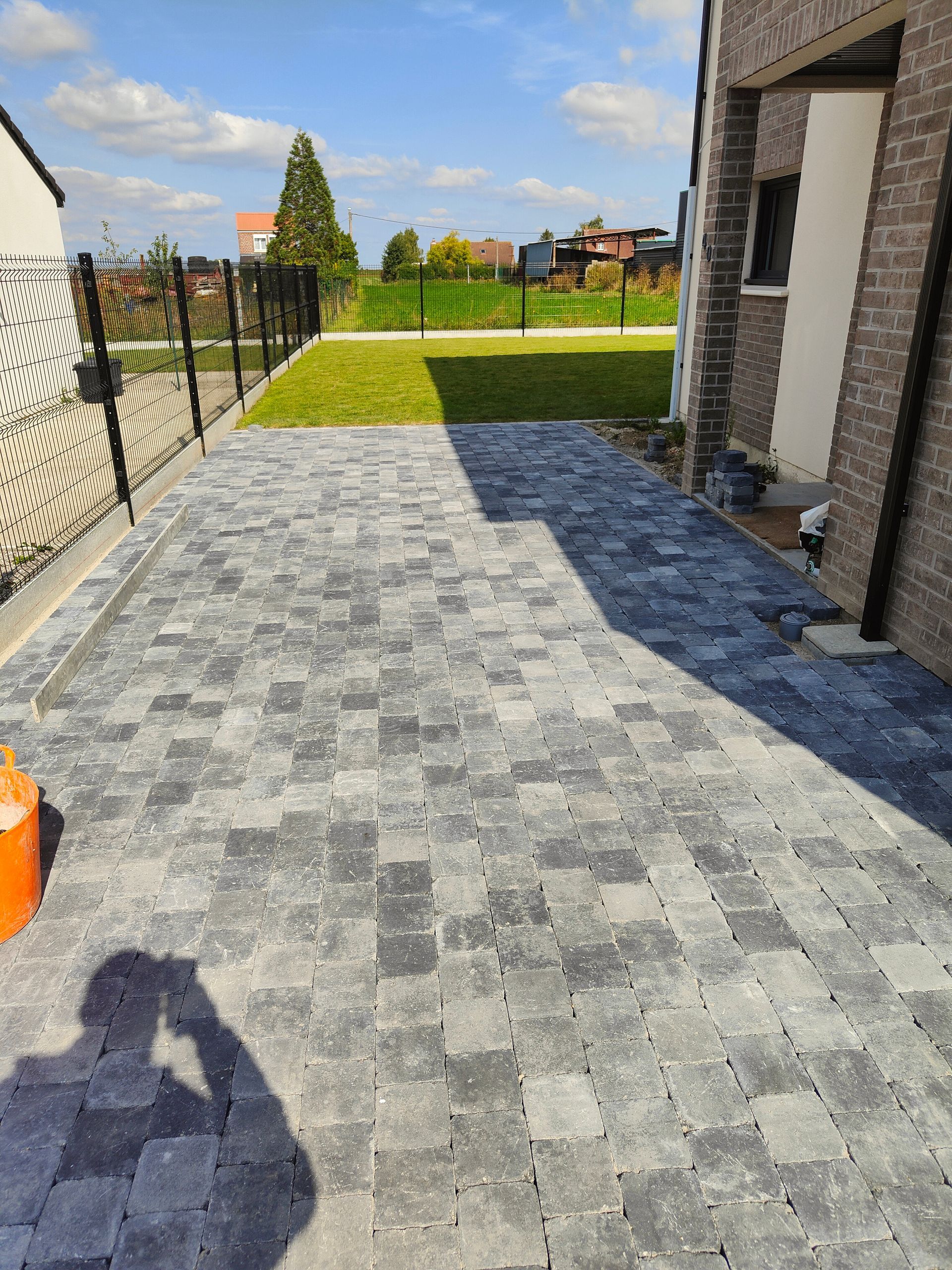 Terrasse arrière avec pavés gris, mur de maison et pelouse verte sous un ciel ensoleillé