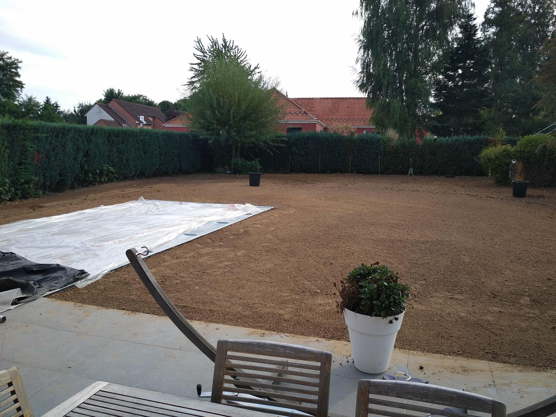 Jardin avec pelouse de gravier, terrasse, haies, arbres et un espace blanc couvert près d'une chaise et d'une plante en pot