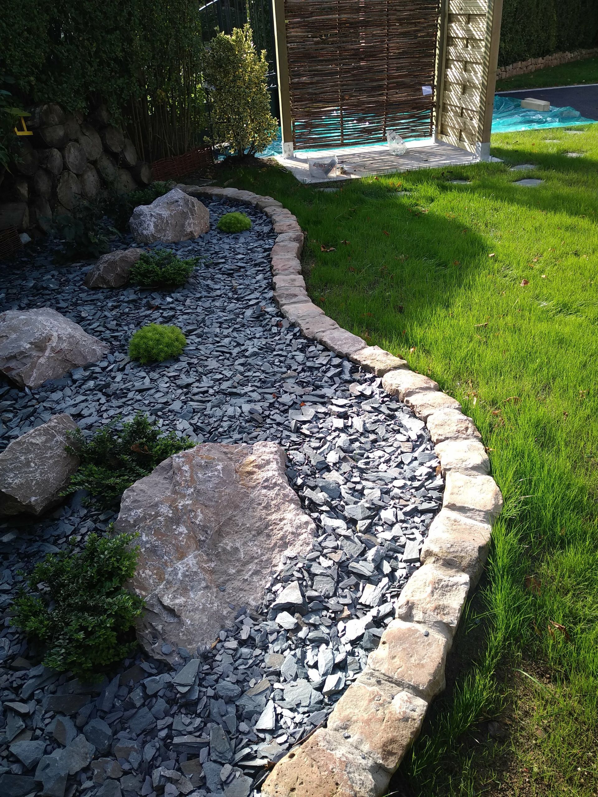Une bordure de pierre incurvée sépare un jardin de rocaille de gravier d'une pelouse d'un vert éclatant, avec une treille en bois et une piscine à l'arrière.