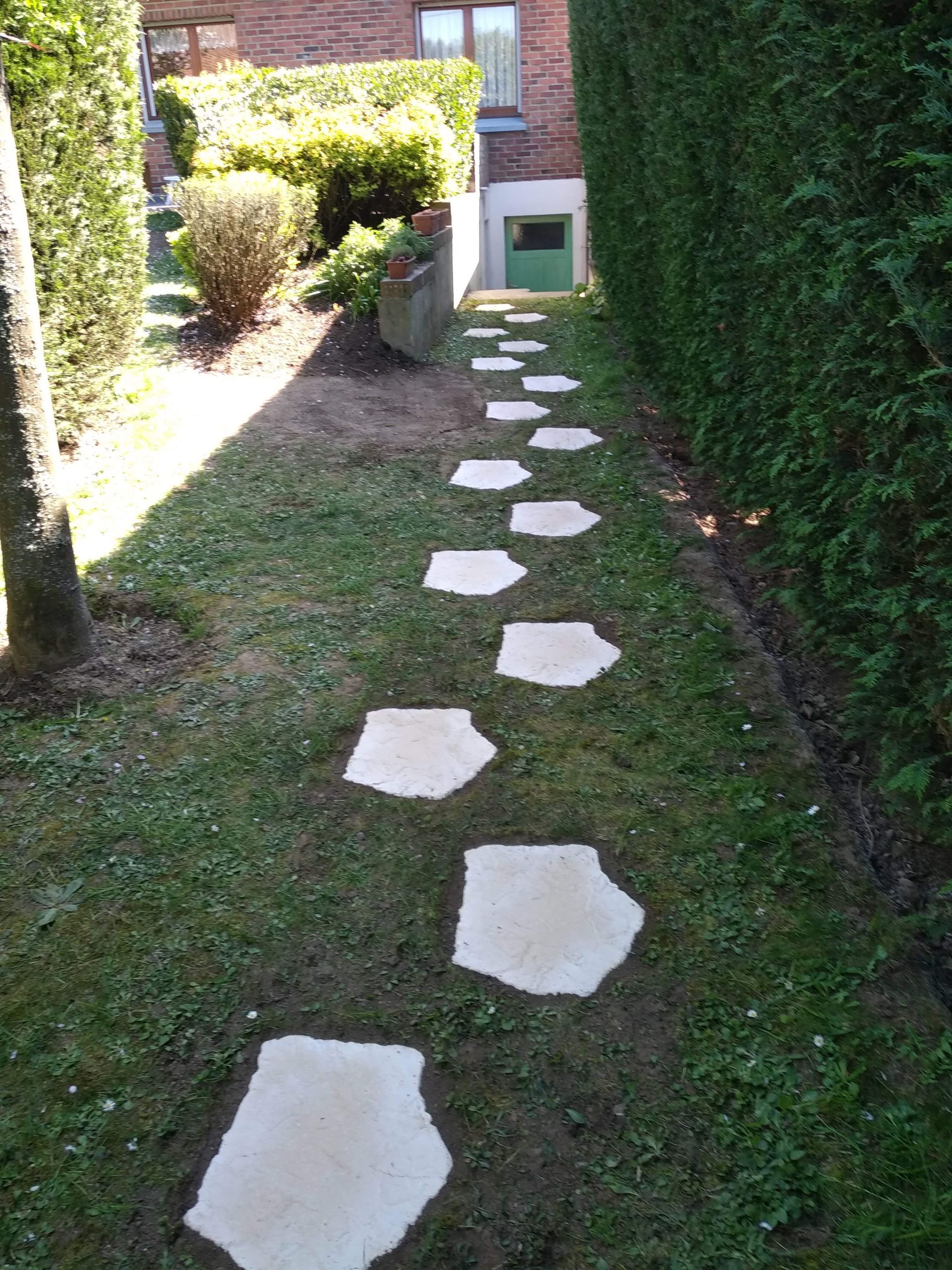 Une allée étroite, agrémentée de dalles blanches, mène à un bâtiment niché entre des haies et des arbres.