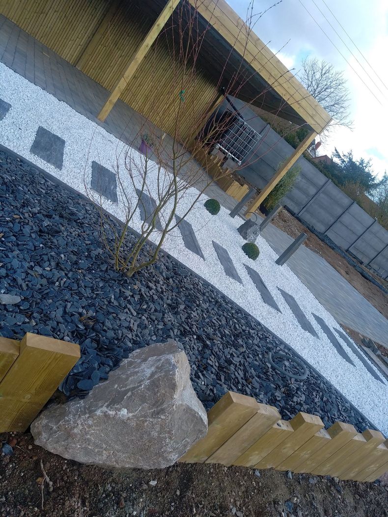 Petit parterre de jardin aménagé avec des graminées ornementales, des pierres sombres et des rochers dans une zone herbeuse près d'un bâtiment.