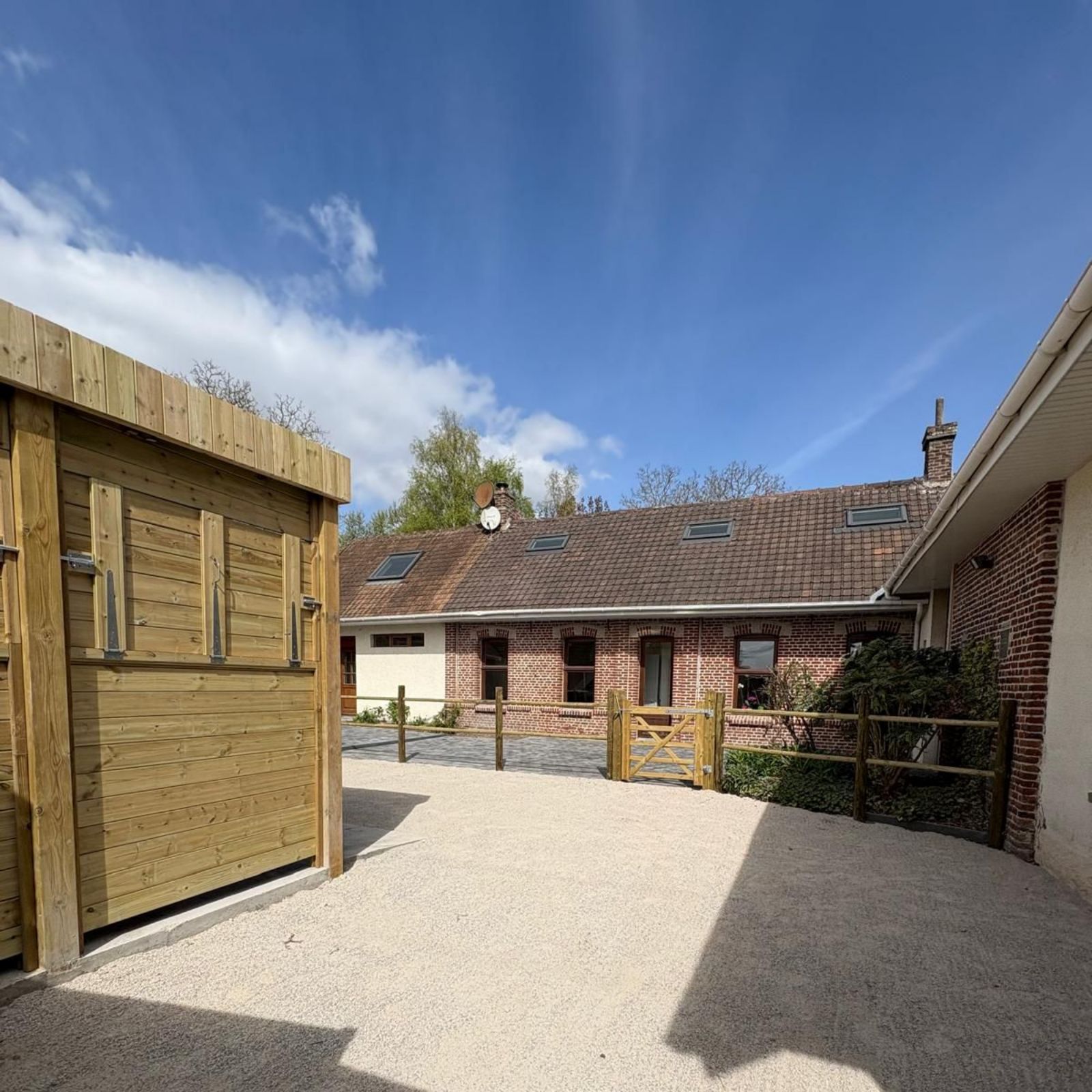 Cour gravillonnée avec abri en bois, clôture et maison en briques sous un ciel bleu