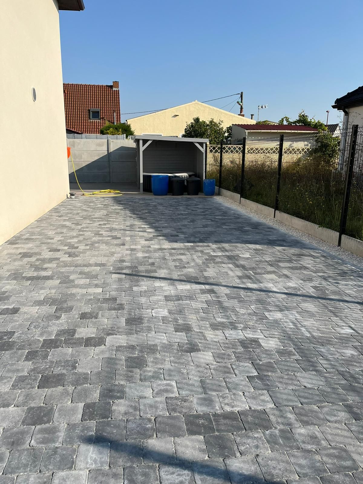Allée pavée menant à un abri à poubelles couvert à côté d'une maison, avec clôture et jardin en arrière-plan.