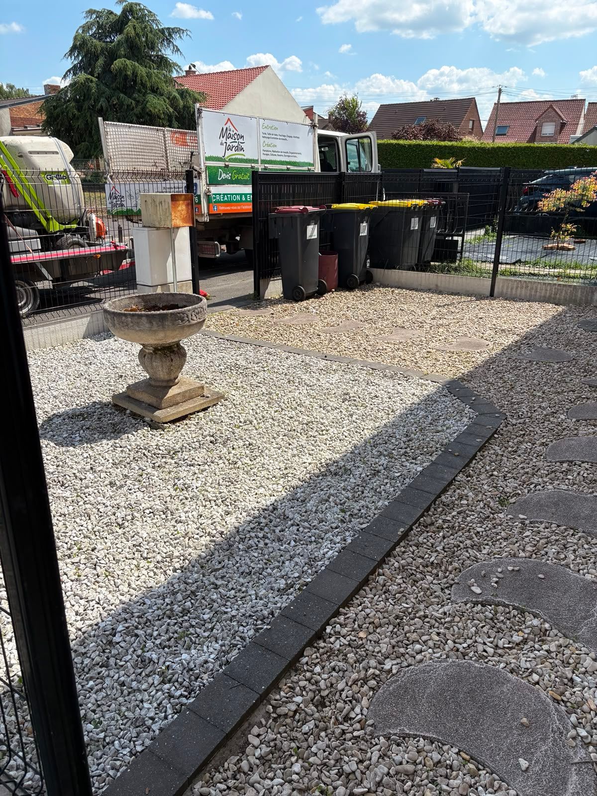 Cour de gravier avec un abreuvoir à oiseaux en pierre, des poubelles noires et une clôture sous un ciel ensoleillé.