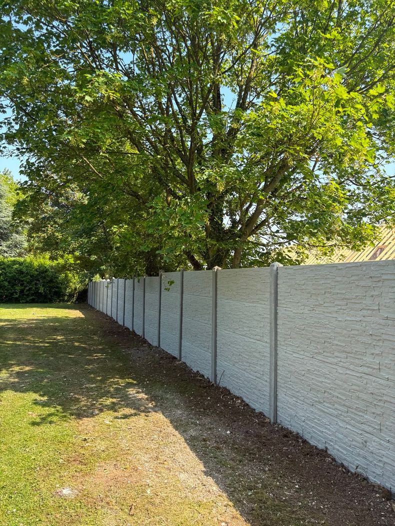 Une longue clôture blanche borde une pelouse surmontée d'un grand arbre baigné de soleil.
