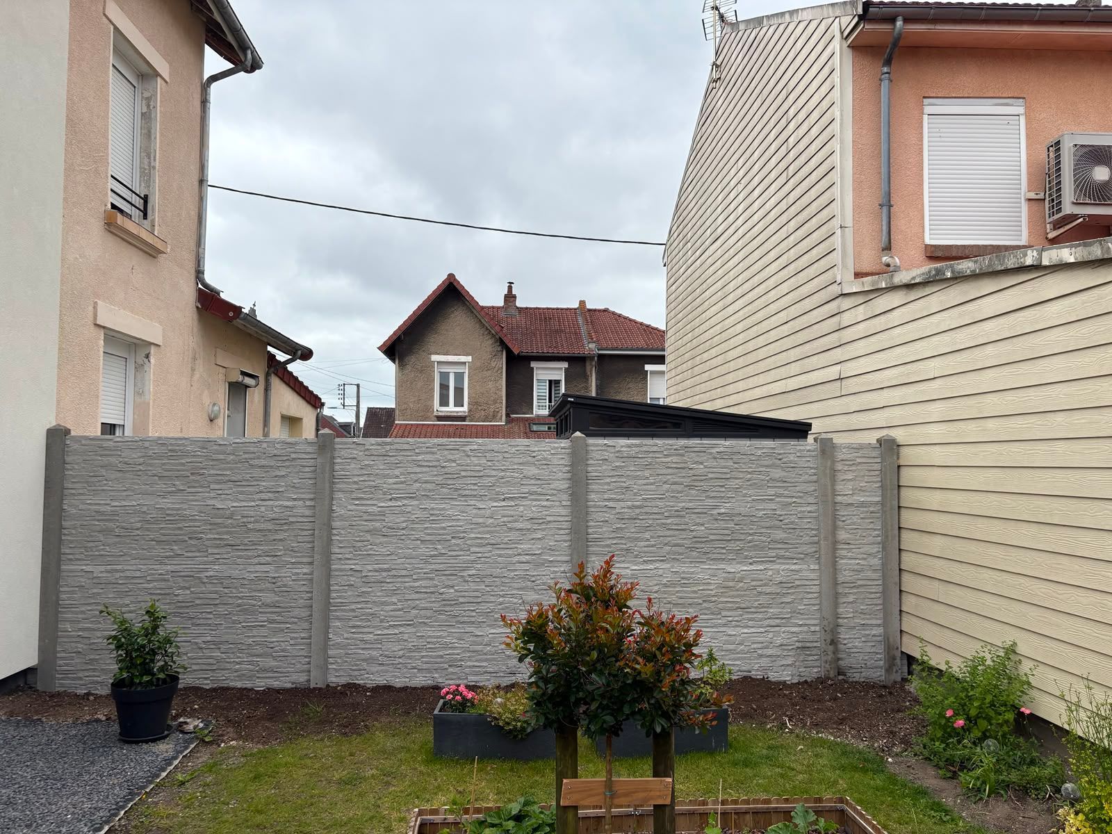 Petit jardin avec muret en pierre, plantes en pot et maisons voisines sous un ciel nuageux