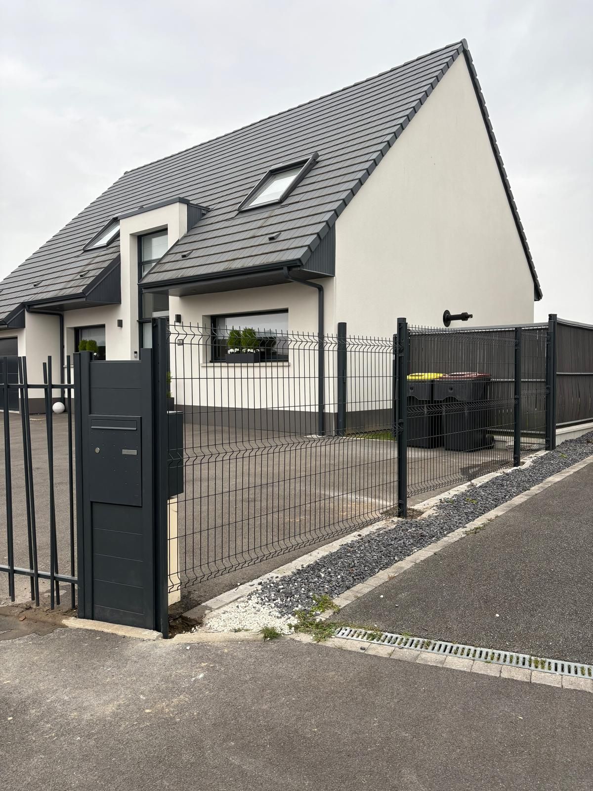 Maison moderne avec un toit gris pentu, un portail noir et une allée clôturée donnant sur une rue pavée.