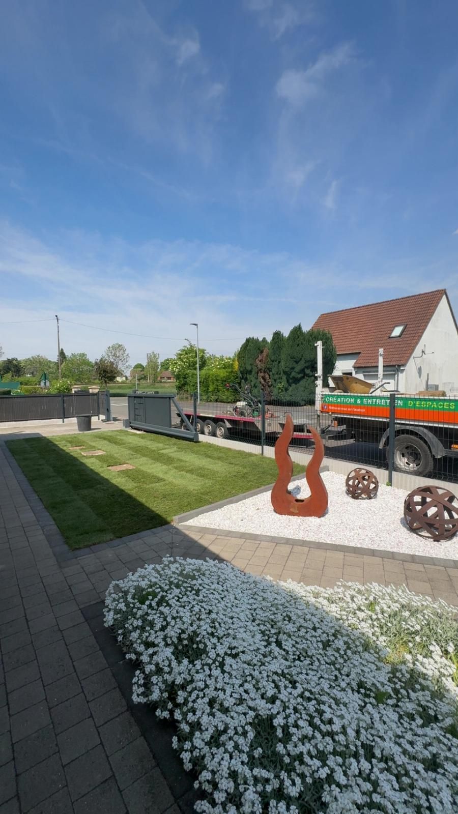 Jardin moderne avec des fleurs blanches, des sculptures en métal, une terrasse pavée et un camion près d'une cour clôturée sous un ciel bleu.
