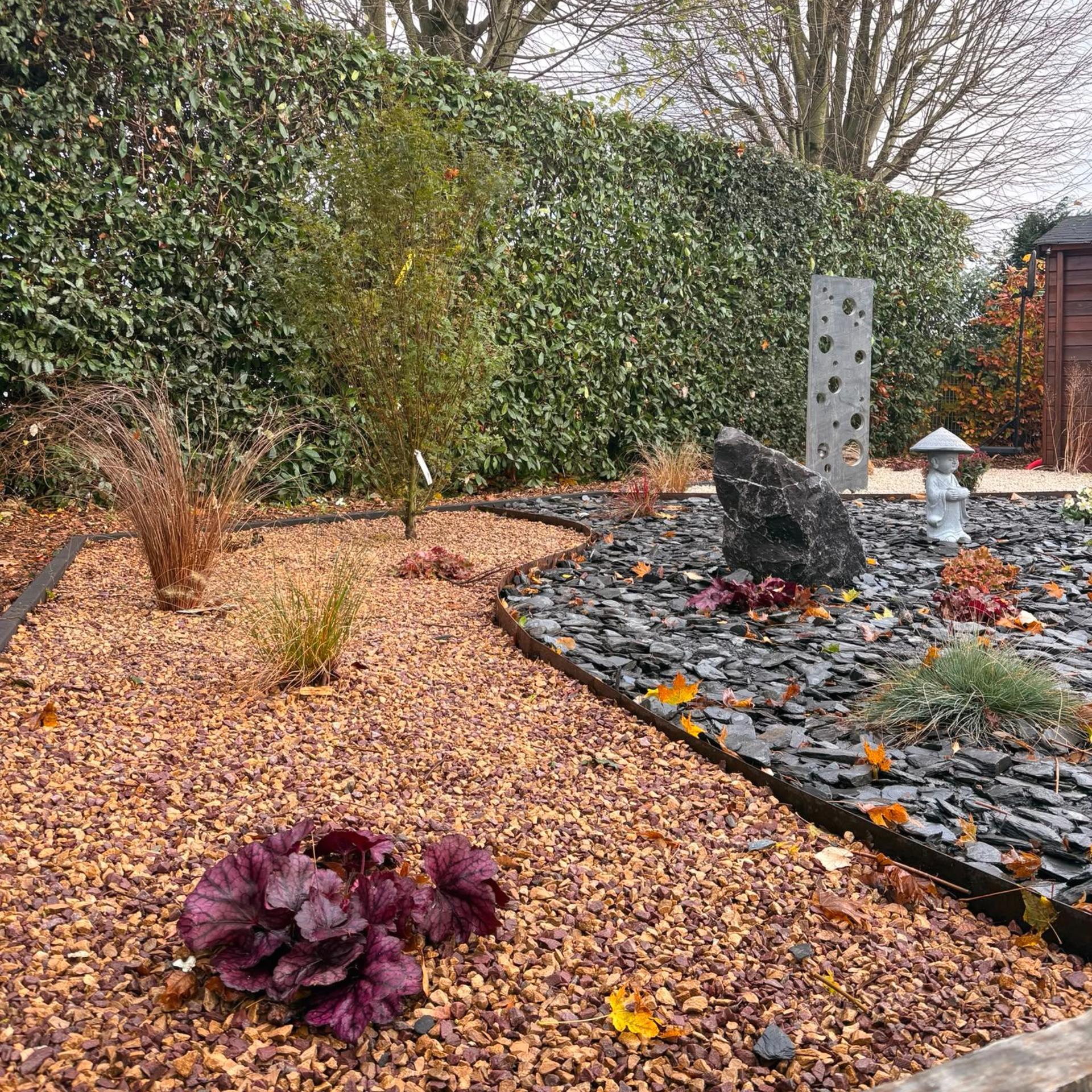 Massif de jardin recouvert de feuilles avec une plante violette, un chemin de pierres, un point d'eau et une haute haie en arrière-plan