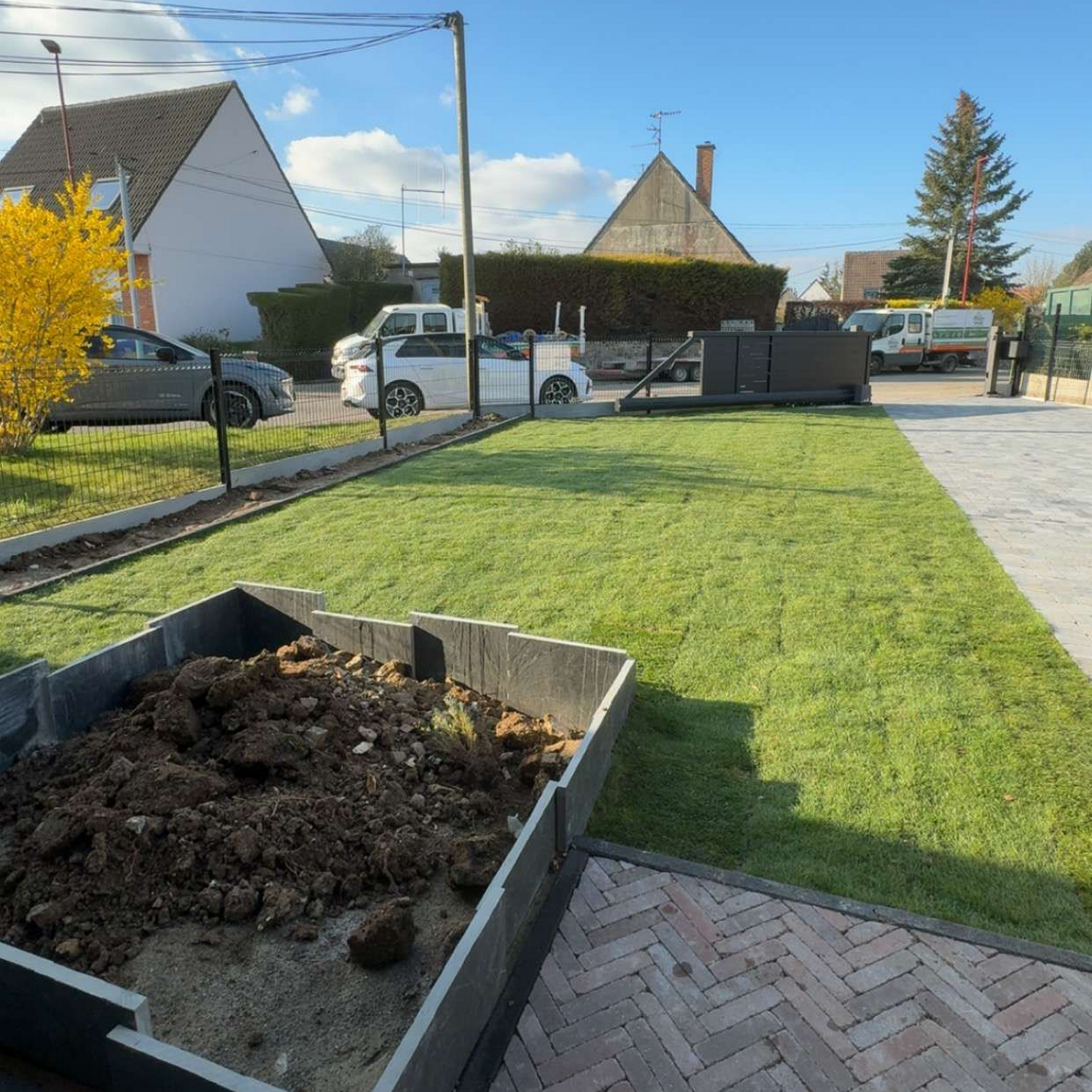 Cour avant avec une brouette remplie de terre, un chemin pavé, une pelouse verte et des maisons en arrière-plan sous un ciel ensoleillé