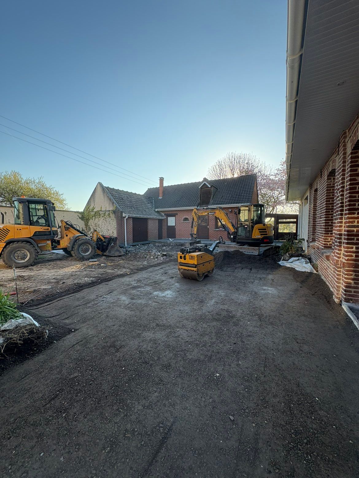 Des engins de chantier et un ouvrier sur un chantier pavé à côté d'un bâtiment en briques sous un ciel dégagé