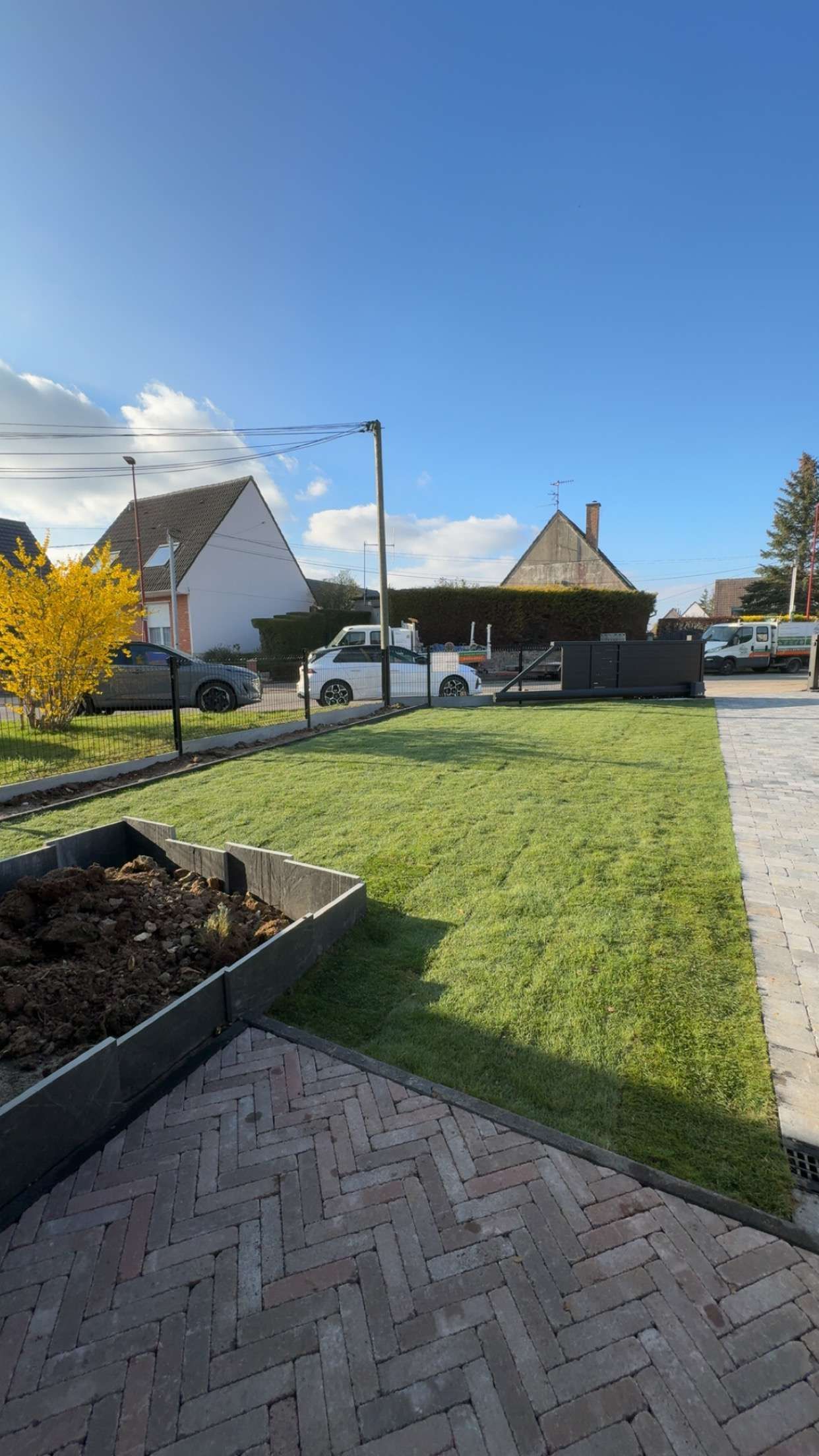 Cour arrière ensoleillée avec pelouse verte, terrasse pavée, potager surélevé et maisons sous un ciel bleu