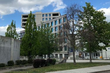 Ein großes Gebäude mit vielen Fenstern ist von Bäumen und Büschen umgeben.