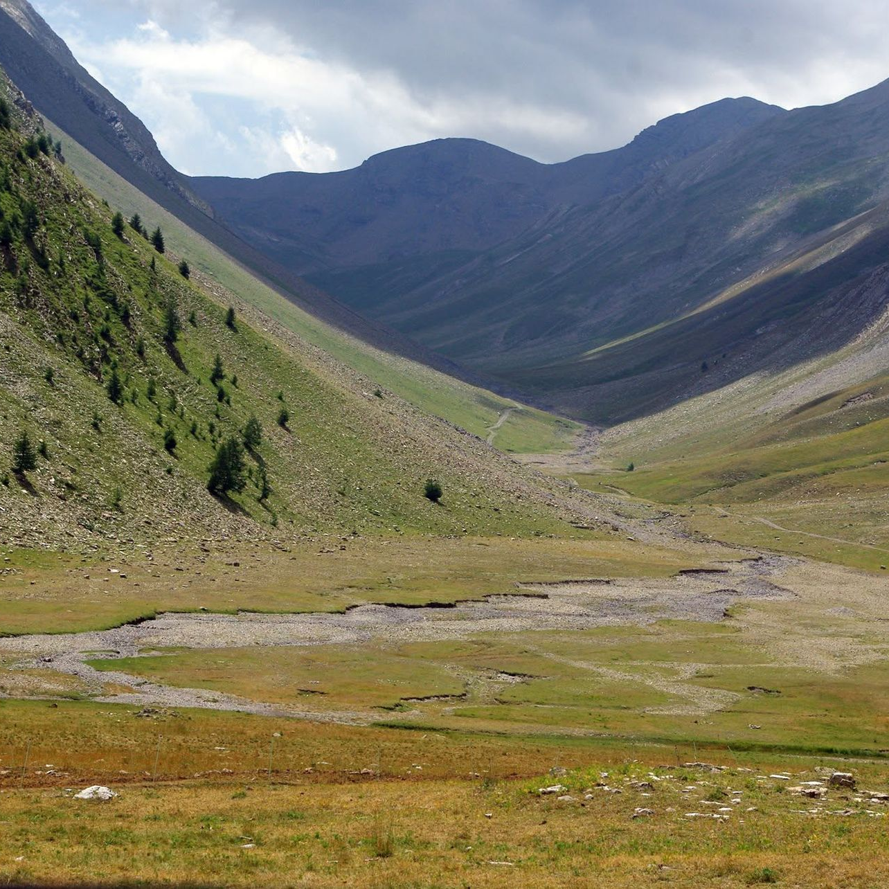 Vallée de montagne aux pentes verdoyantes, un ruisseau sinueux et des sommets lointains.