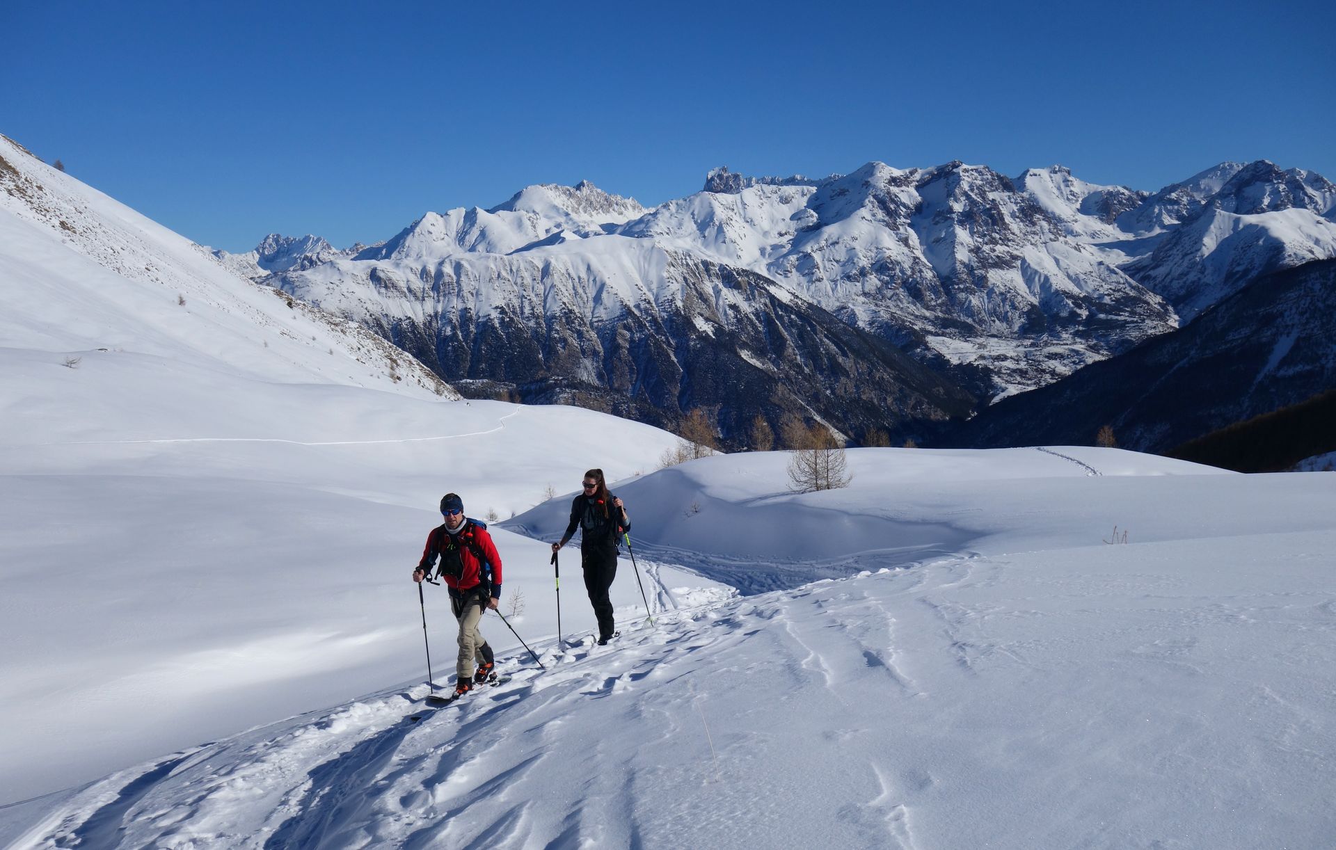Deux randonneurs traversent un versant enneigé d'une montagne, avec des sommets en arrière-plan.