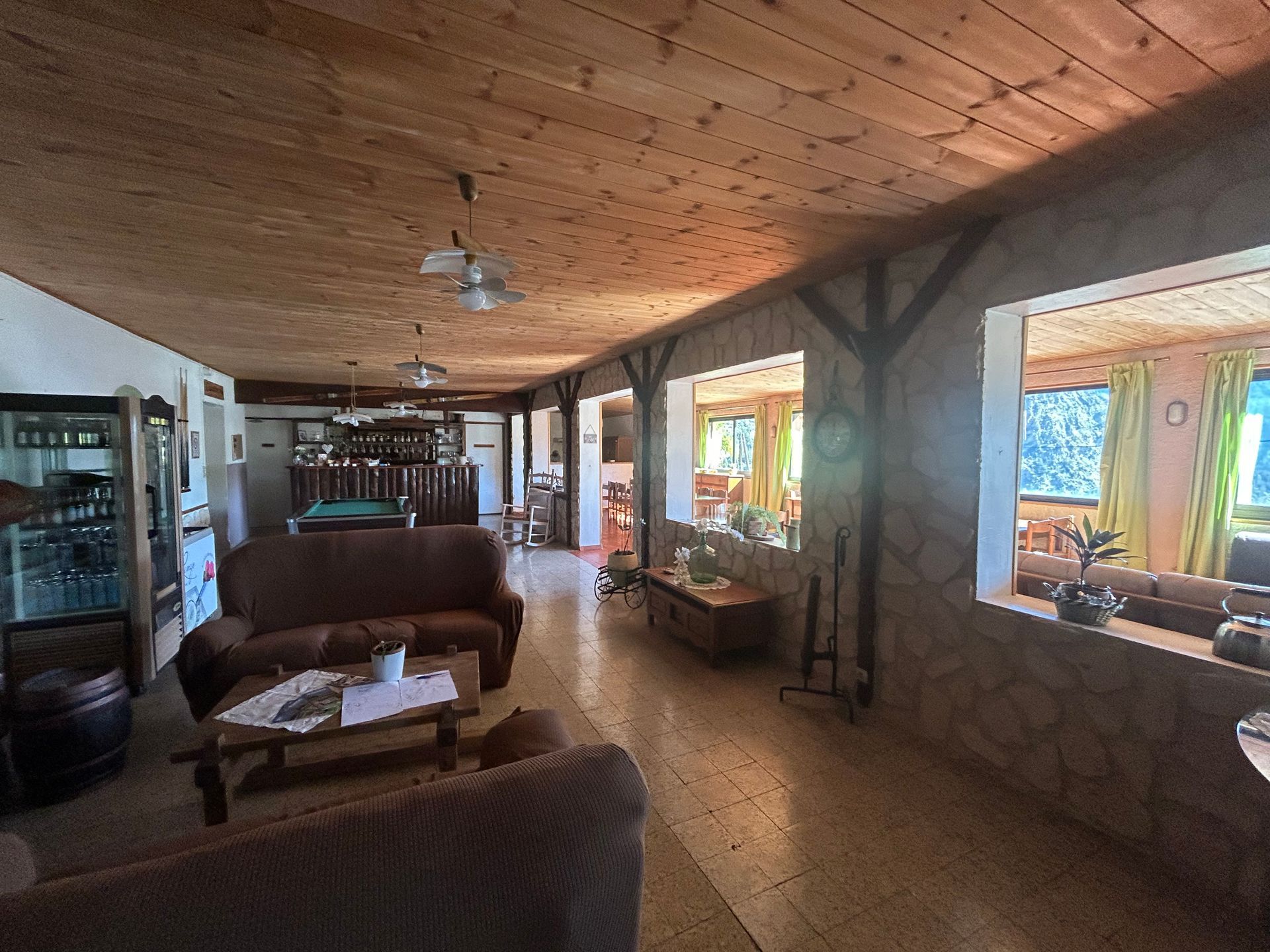 Salon rustique avec plafond en bois, murs en pierre, canapés et grandes fenêtres.