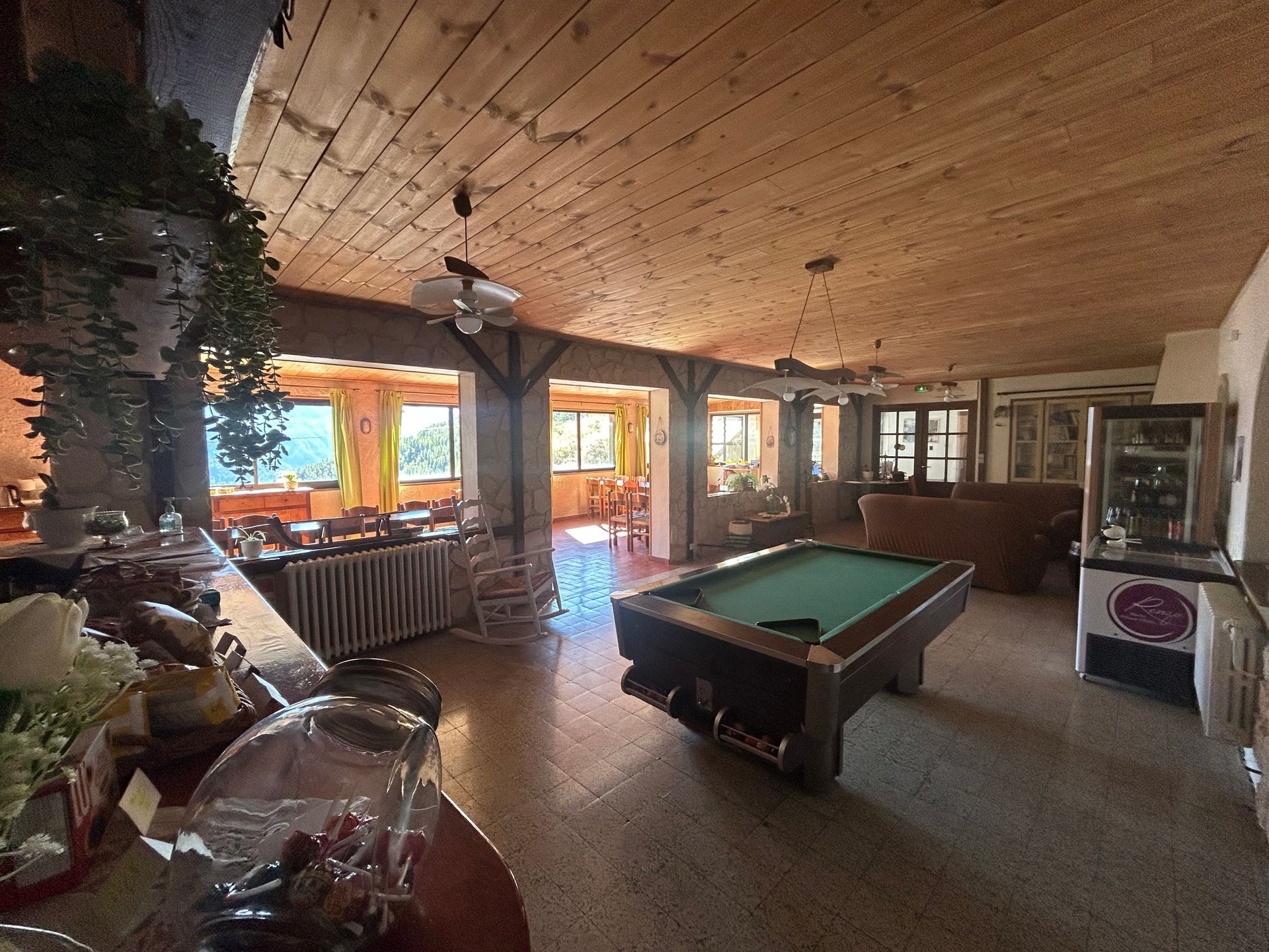 Salon rustique avec table de billard, plafond lambrissé, cheminée et grandes fenêtres.