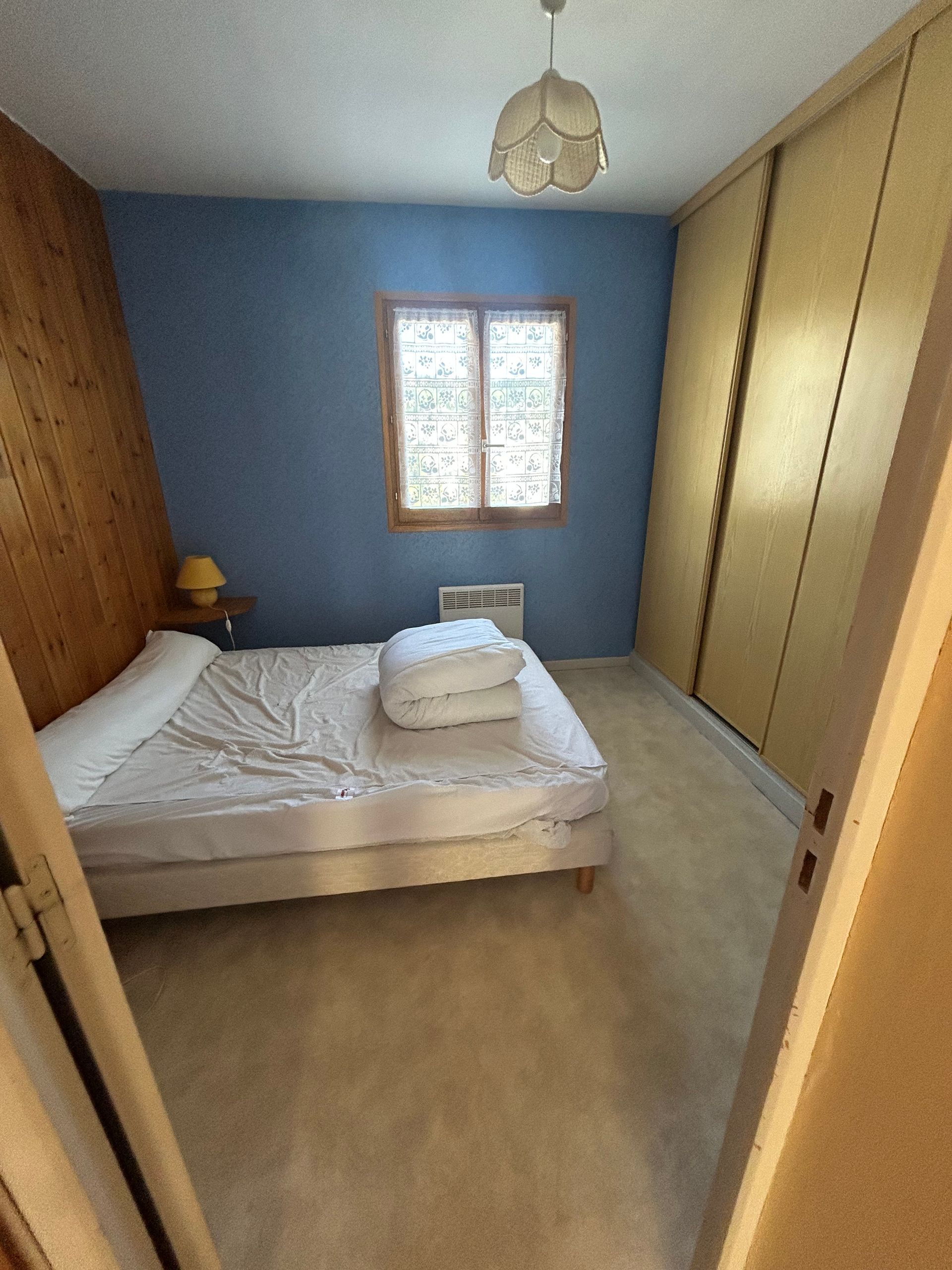 Petite chambre avec murs bleus, un lit, une fenêtre et une grande armoire encastrée.