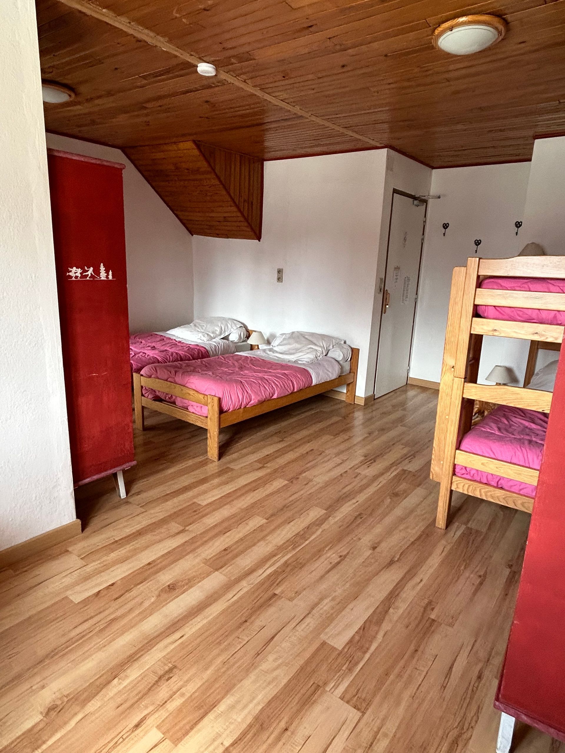 Petite chambre mansardée avec plafond en bois, lit rose, lit superposé, armoire rouge et parquet.